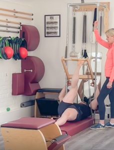 Man lying on mat with right foot on wooden bar performing hamstring stretch. Teacher assisting.
